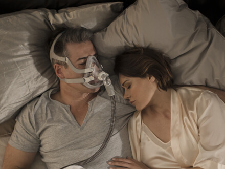 Casal dorme numa cama. Homem utiliza a máscara facial Versátil AirFit F20