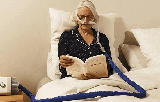 Mulher idosa deitada na sua cama conectada ao seu dispositivo faz terapia enquanto lê um livro
