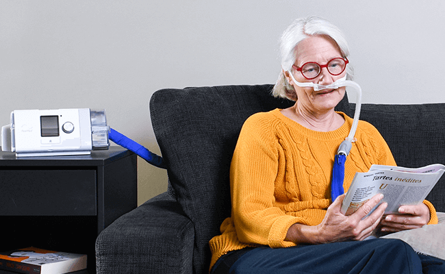 Senhora de idade usa o seu dispositivo de terapia enquanto descansa no sofá e lê uma revista